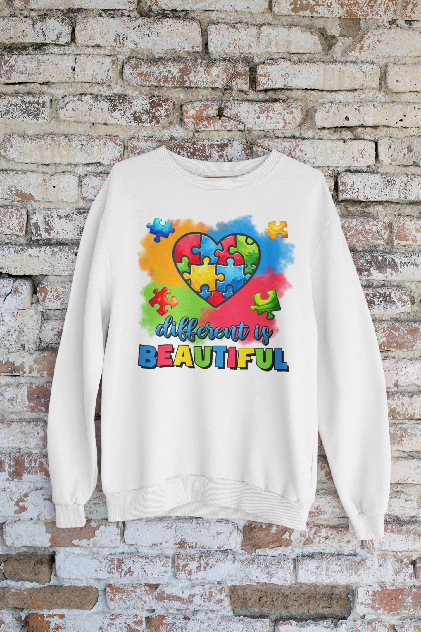 White sweatshirt with colorful heart design and 'different is beautiful' text against a brick wall.