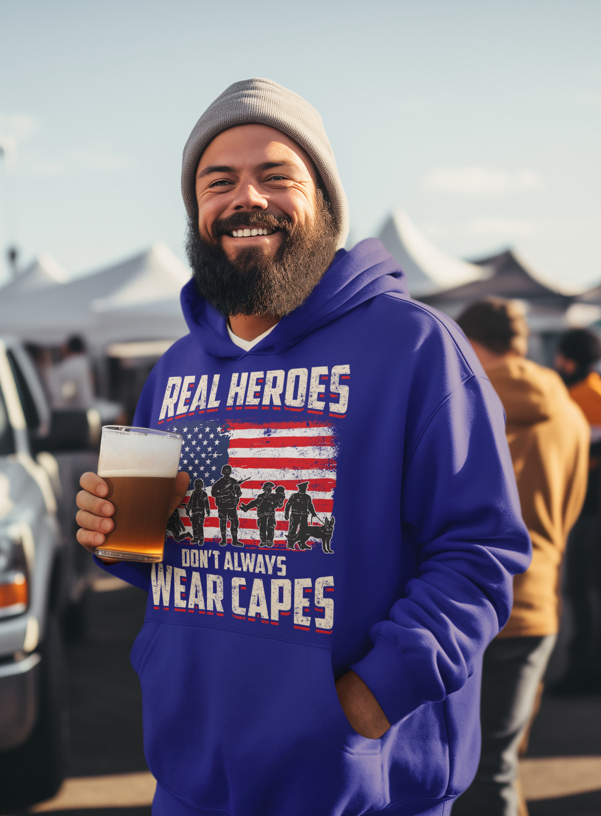 Man wearing a blue hoodie with a graphic and text, holding a beer at an outdoor event.