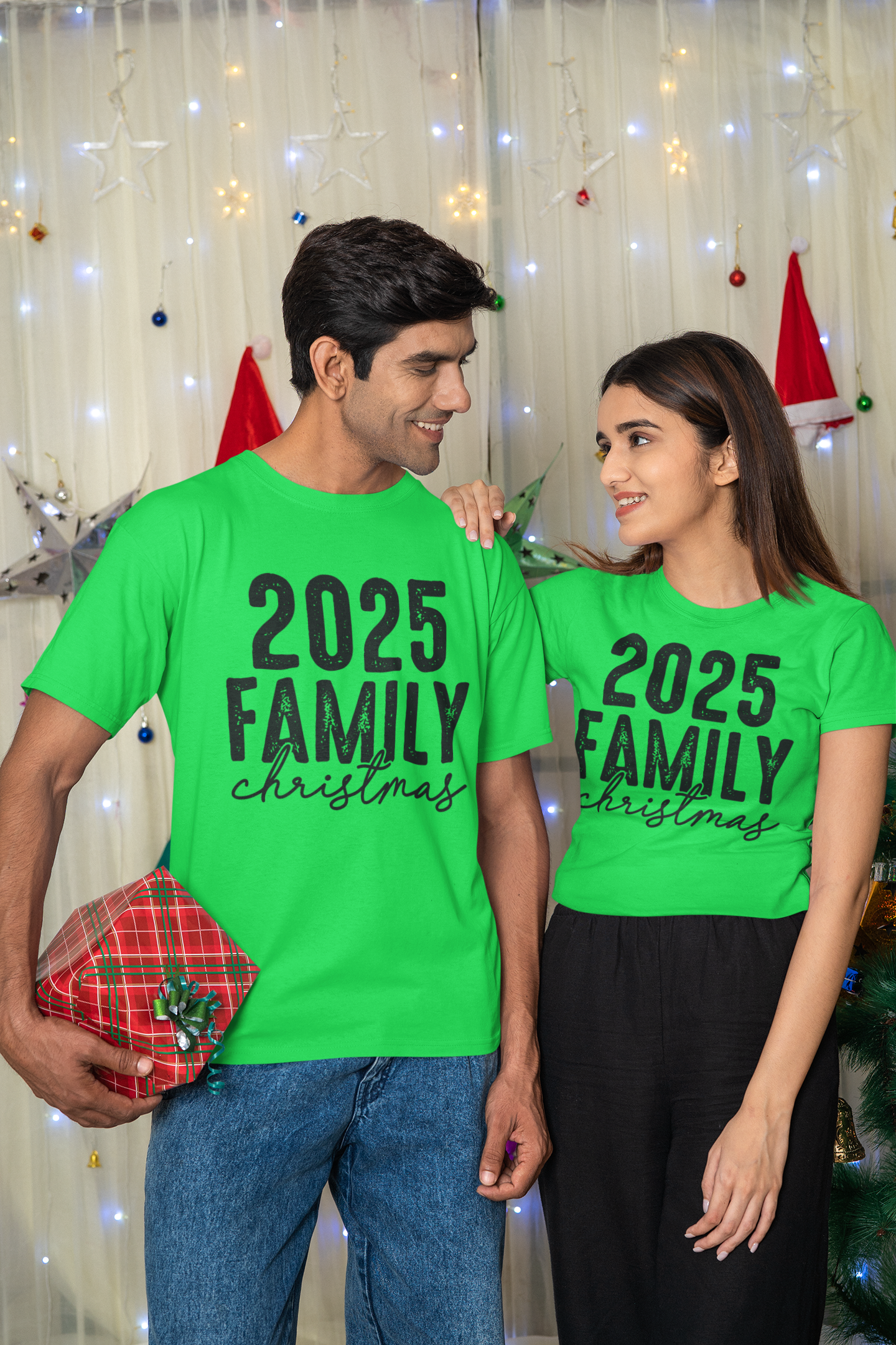 Two people wearing green t-shirts with '2025 Family Christmas' text, standing in front of a festive background.
