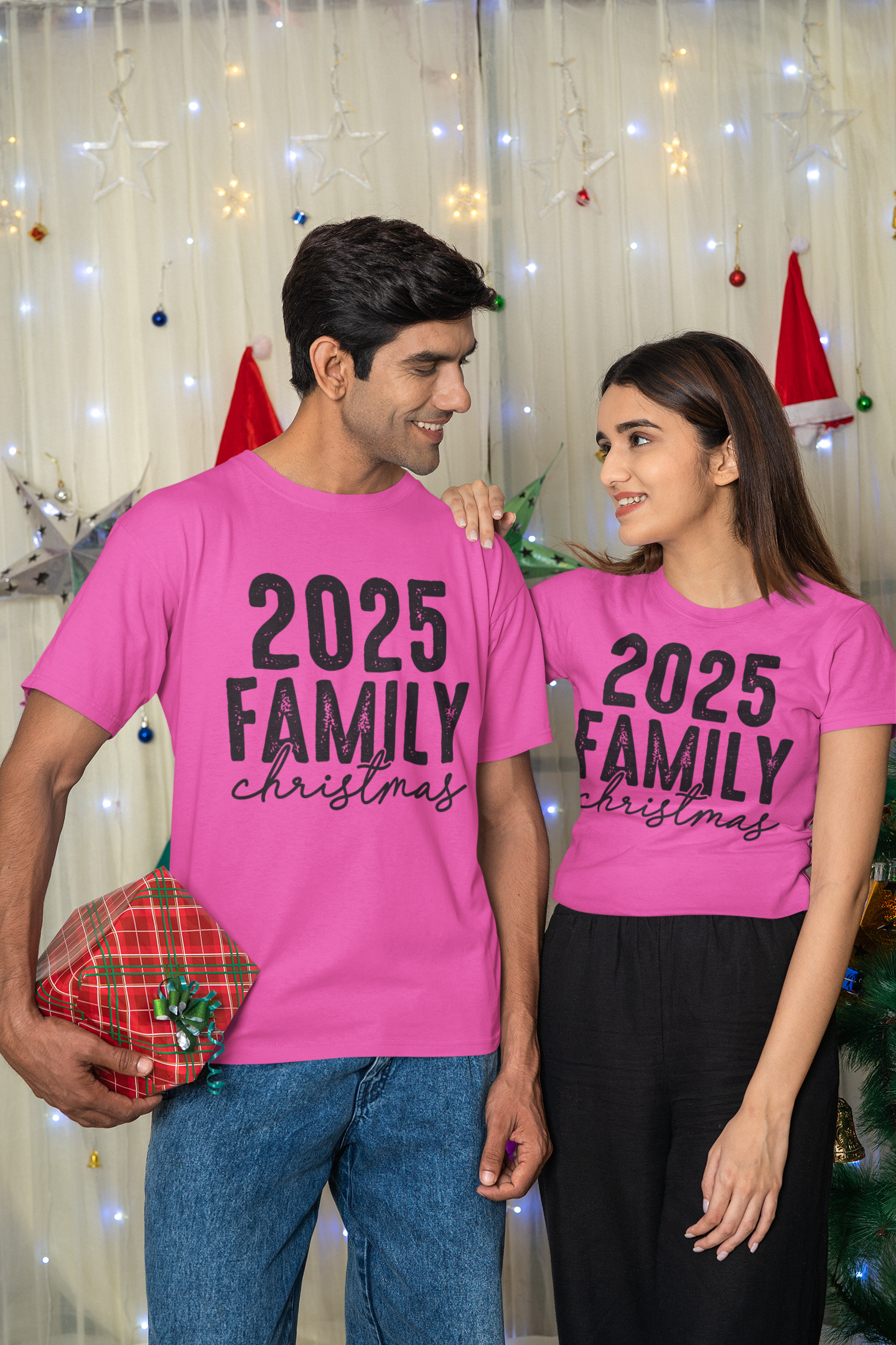 Two people wearing pink '2025 Family Christmas' shirts in front of a festive background.