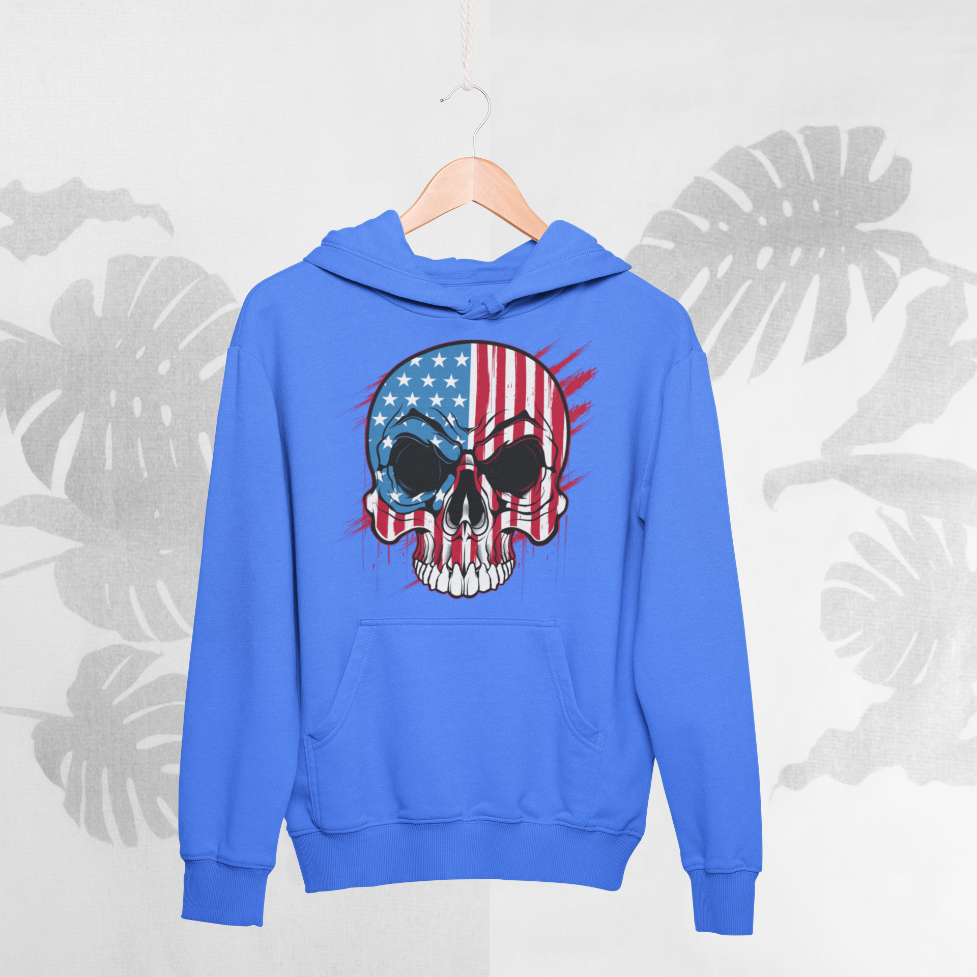Blue hoodie with a skull and American flag design on a hanger against a light gray background with leaf patterns.