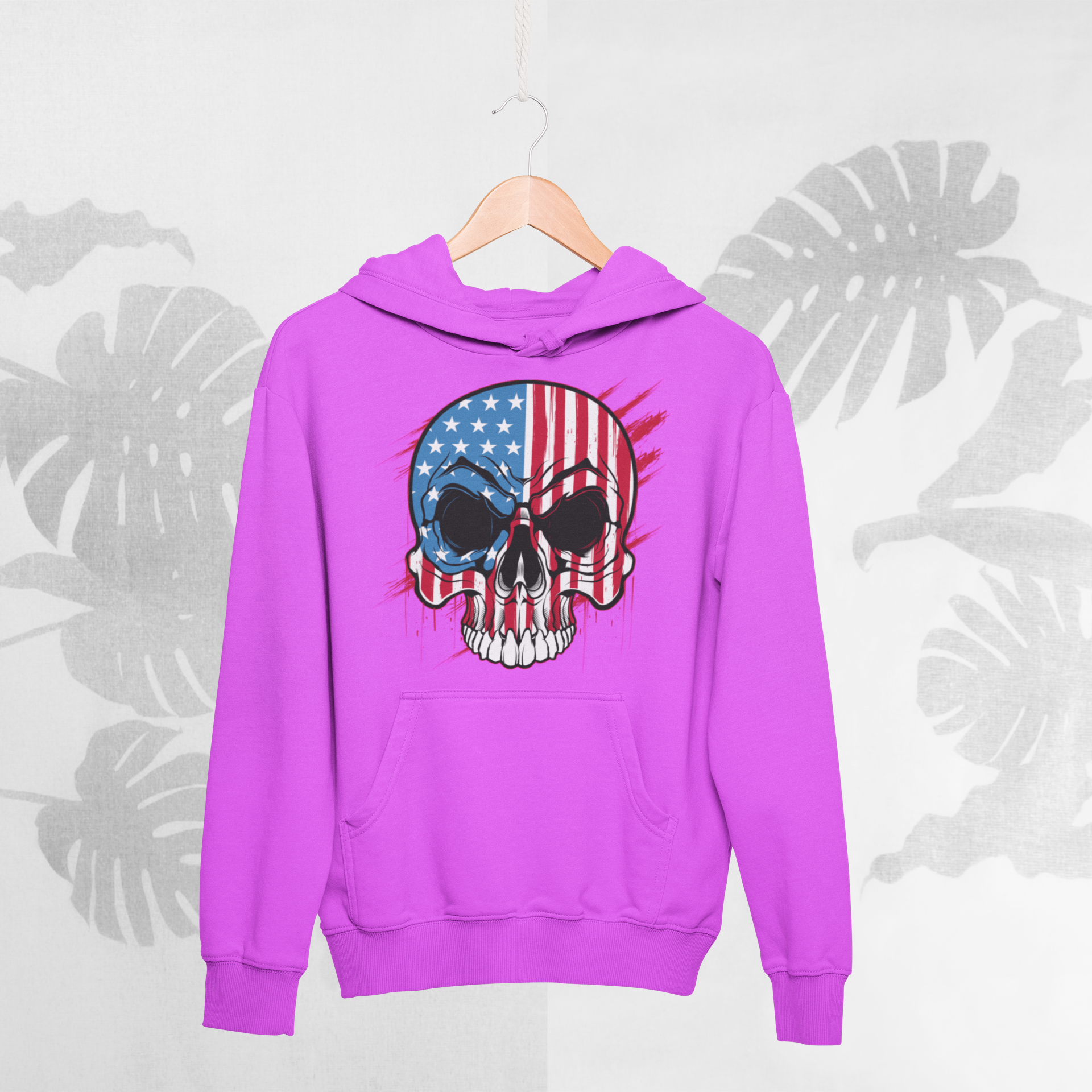 Pink hoodie with a skull and American flag design on a hanger against a light gray background with leaf patterns.