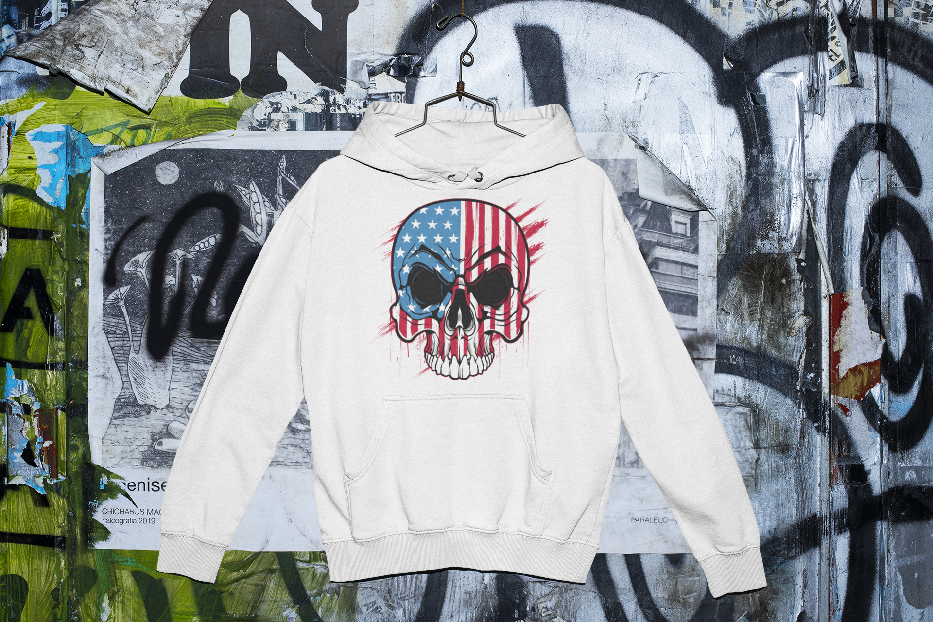 White hoodie with skull and American flag design on a graffiti-covered wall.