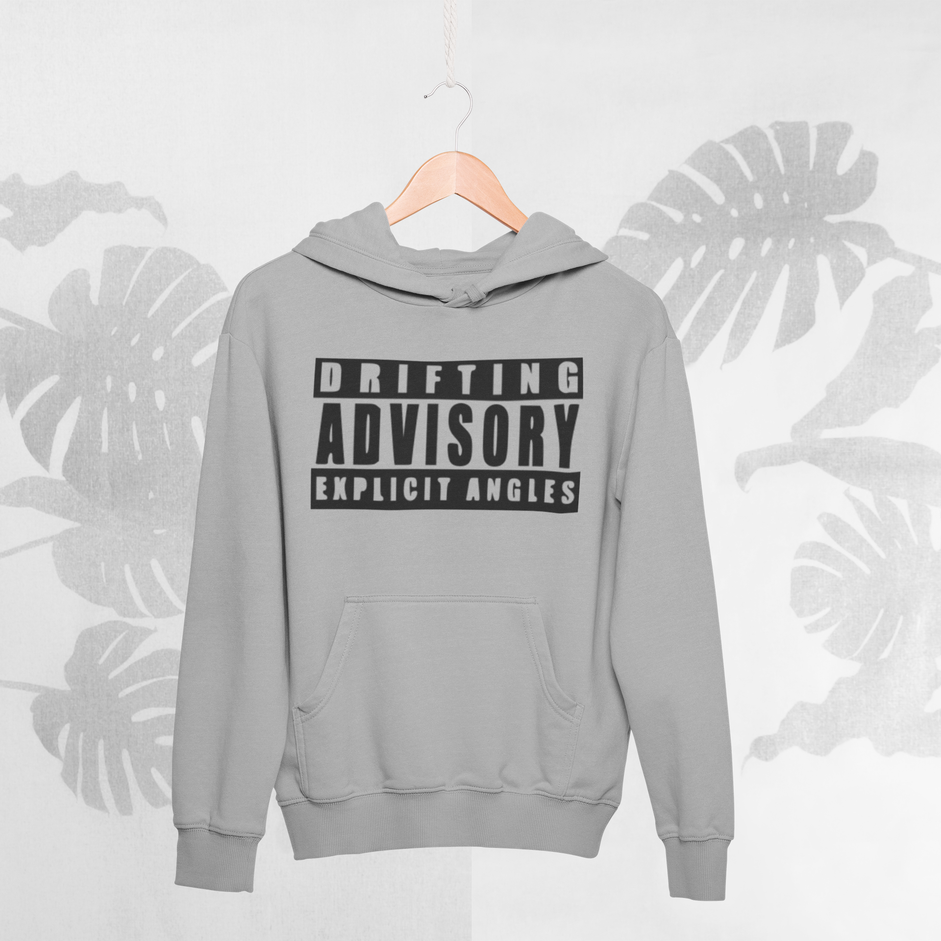 Gray hoodie with 'Drifting Advisory Explicit Angles' text on a hanger against a light gray background with leaf patterns.