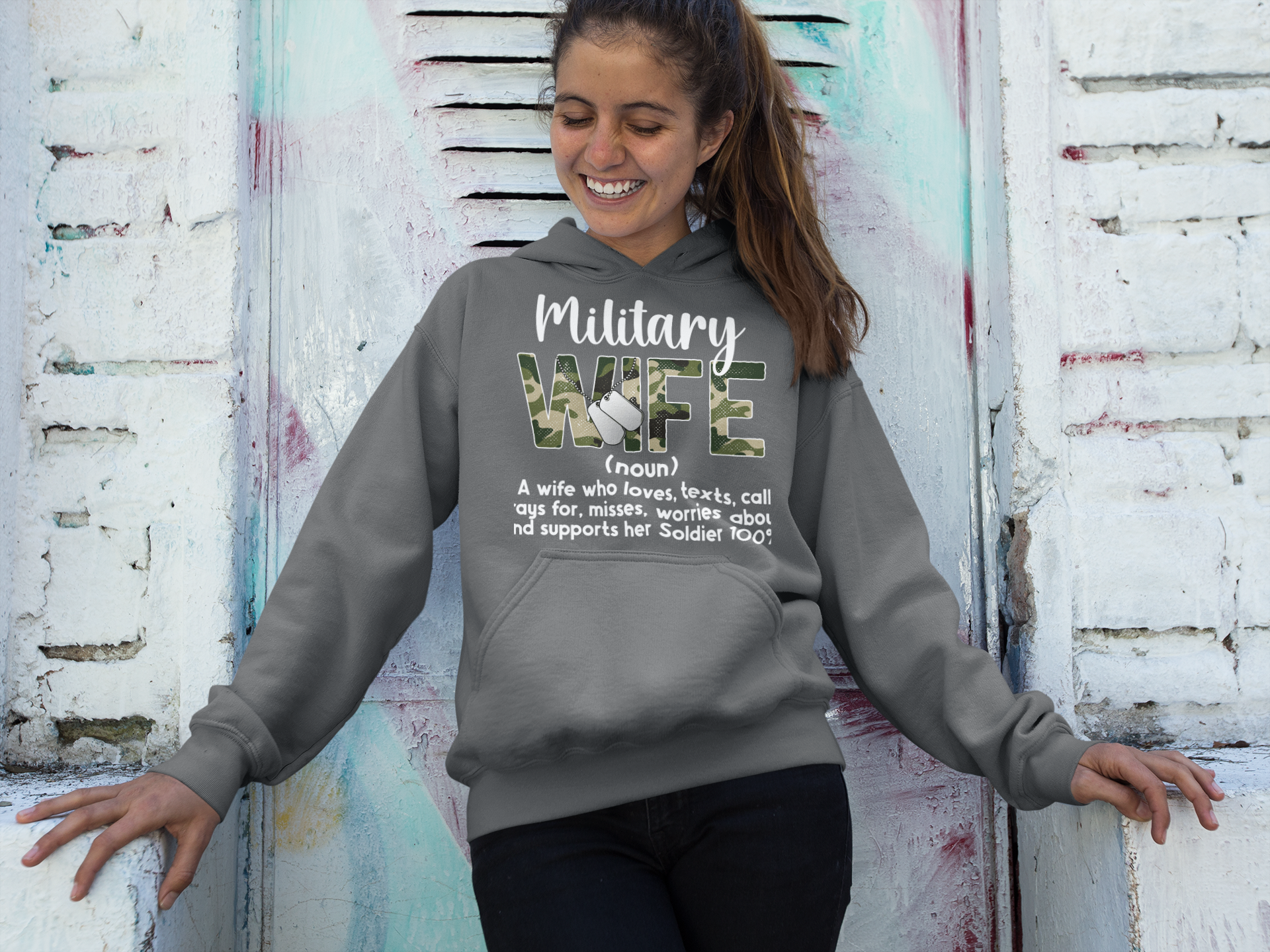 Woman wearing a gray hoodie with 'Military Wife' text and definition against a textured wall.