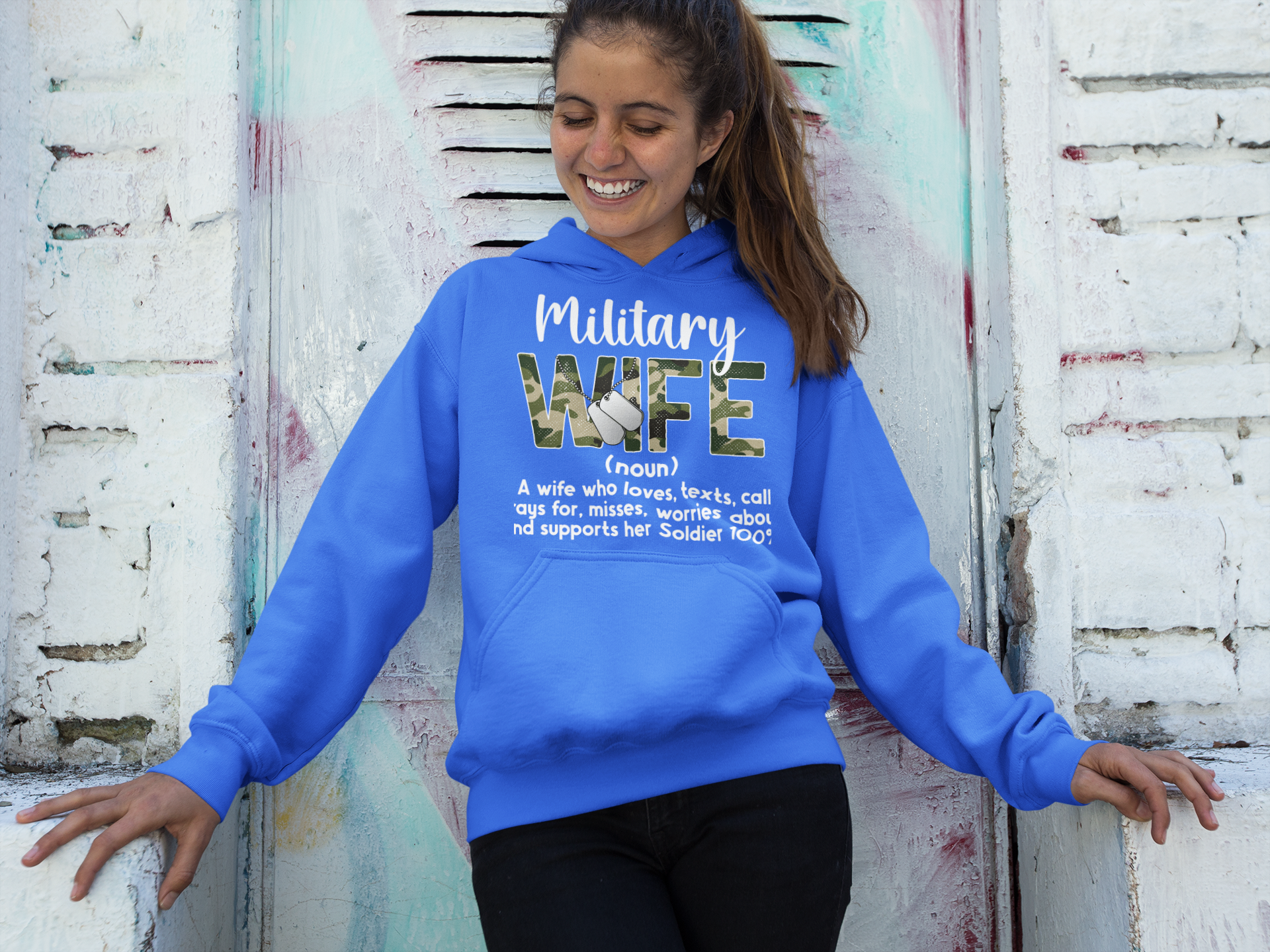 Woman wearing a blue hoodie with 'Military Wife' text against a textured wall.